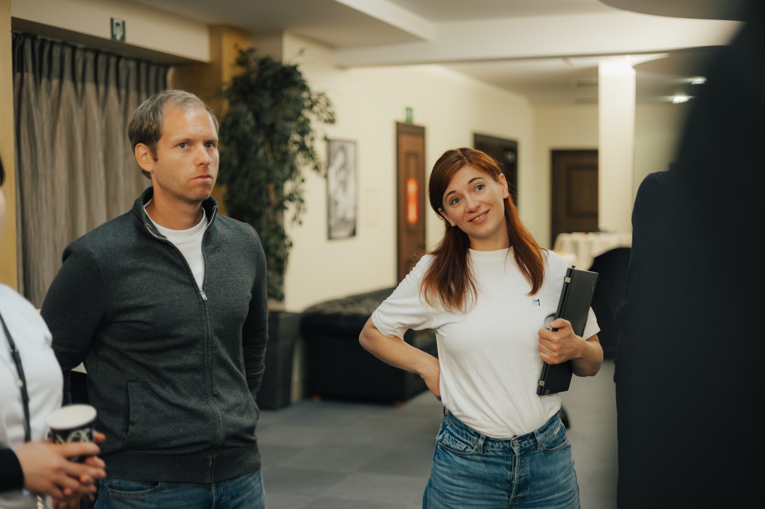 NEO.cation 2025 Zwei Personen stehen in einem Meetingraum und die linke lächelt während sie im Gespräch ist. Rechts daneben steht eine Person die gespannt zuhört
